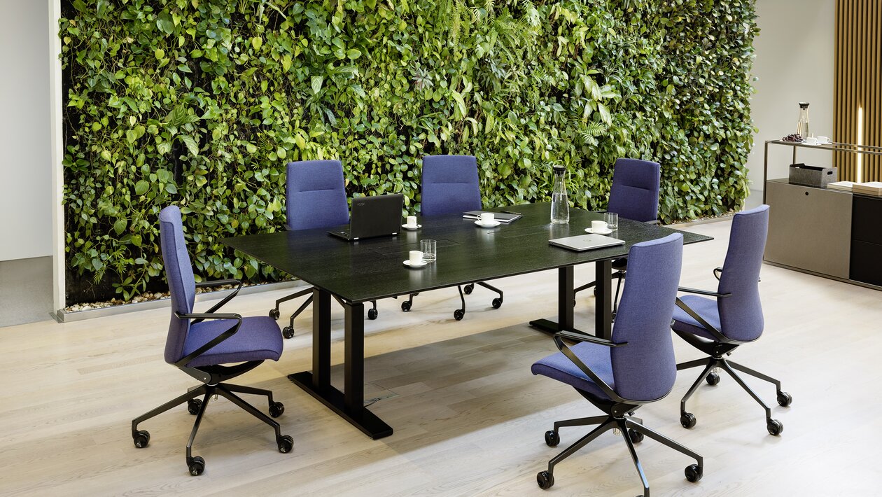 Dunkler Konferenztisch mit violetten Konferenzstühlen vor einer bepflanzten Wand. 
