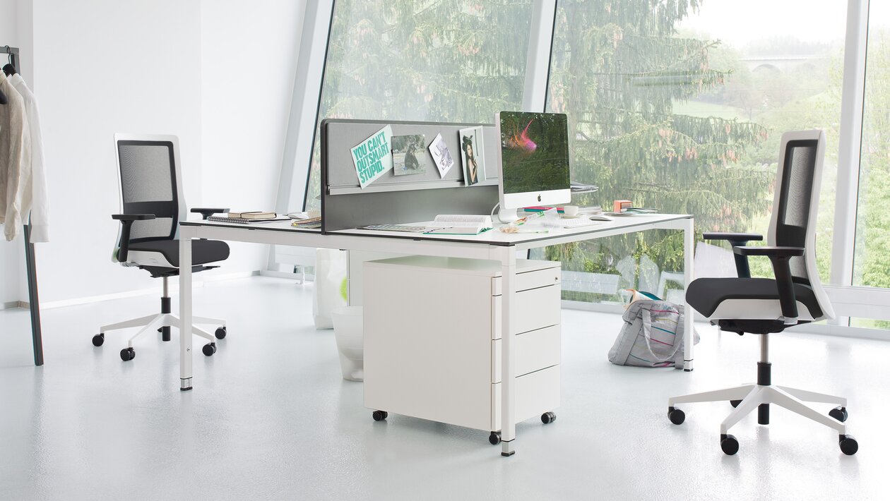 Helles Büro mit weißem Schreibtisch und weiß-schwarzen Bürostühlen.