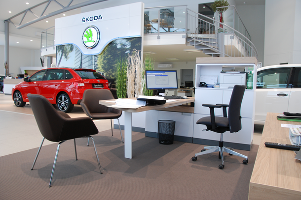 Skoda Braune Konferenzstühle vor einem weißen Schreibtisch mit schwarzem Drehstuhl. Im Hintergrund ist ein rotes Auto zu sehen.