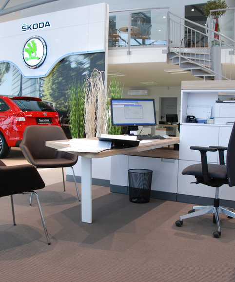 Skoda Braune Konferenzstühle vor einem weißen Schreibtisch mit schwarzem Drehstuhl. Im Hintergrund ist ein rotes Auto zu sehen.