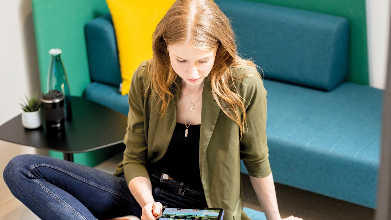 Person sitzt auf einem Polstermöbel und arbeitet auf einem Tablet. 