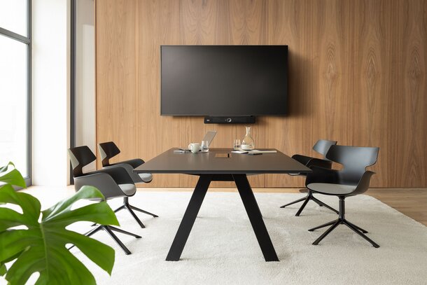 Raum mit einem Fernseher, einem großem Tisch und schwarzen Schalenstühlen mit einer grauen Polsterung und 4 Beinen