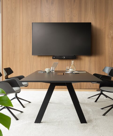 Raum mit einem Fernseher, einem großem Tisch und schwarzen Schalenstühlen mit einer grauen Polsterung und 4 Beinen