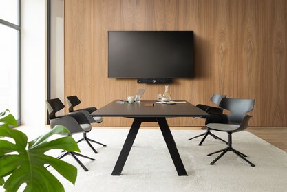 Raum mit einem Fernseher, einem großem Tisch und schwarzen Schalenstühlen mit einer grauen Polsterung und 4 Beinen