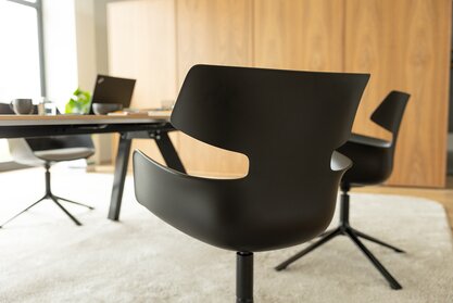 Raum mit einem Holztisch, einem grauen Teppich und schwarzen Schalenstühlen mit grauem Polster und 4 Beinen