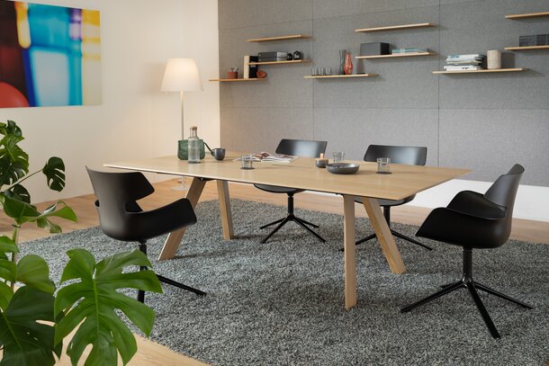Raum mit einem Holztisch, einem grauen Teppich und schwarzen Schalenstühlen mit grauem Polster und 4 Beinen