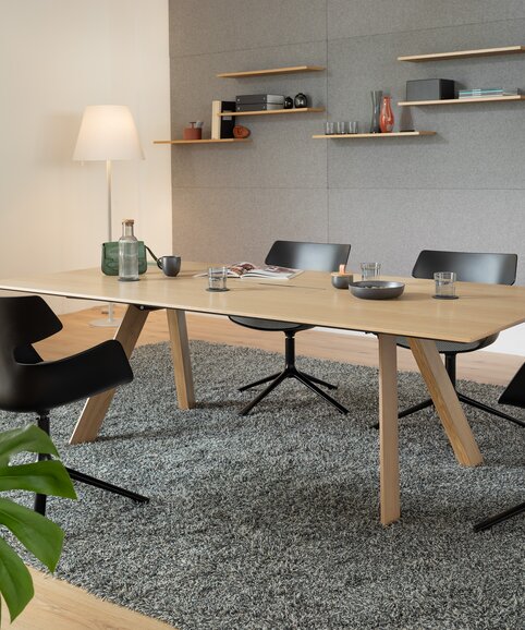 Raum mit einem Holztisch, einem grauen Teppich und schwarzen Schalenstühlen mit grauem Polster und 4 Beinen