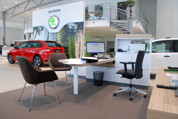 Des chaises de conférence marron devant un bureau blanc avec une chaise tournante noire, avec une voiture rouge à l’arrière-plan.