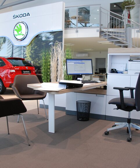 Des chaises de conférence marron devant un bureau blanc avec une chaise tournante noire, avec une voiture rouge à l’arrière-plan.