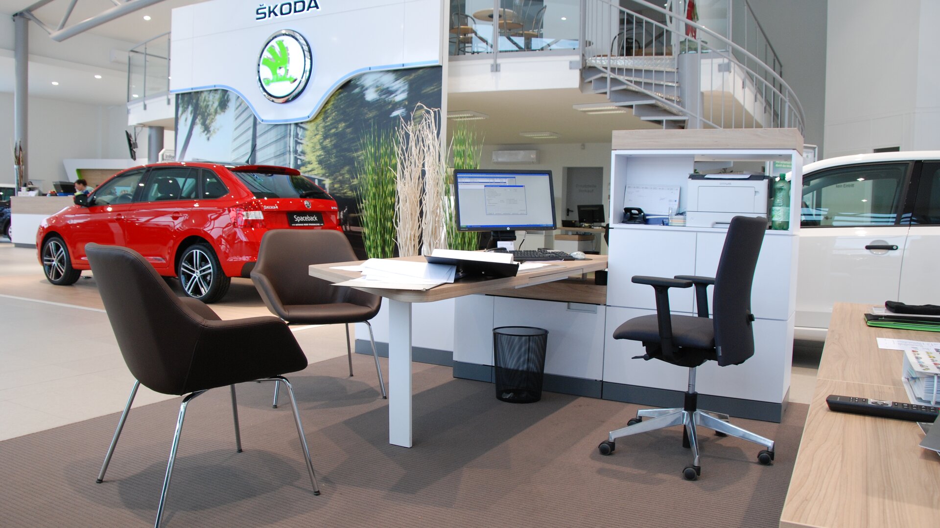 Braune Konferenzstühle vor einem weißen Schreibtisch mit schwarzem Drehstuhl. Im Hintergrund ist ein rotes Auto zu sehen.