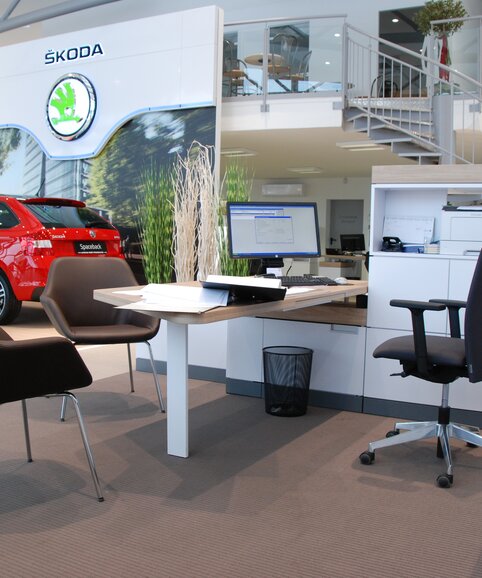 Bruine conferentiestoelen voor een wit bureau met een zwarte bureaustoel. Op de achtergrond is een rode auto te zien