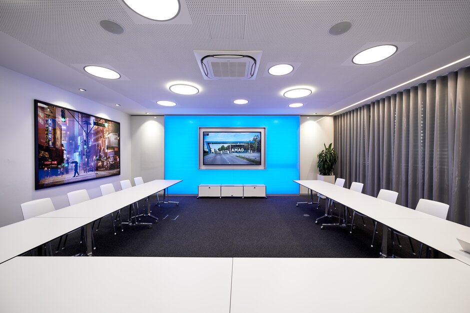 Großer Konferenzraum mit Bildschirm, weißen Tischen und weißen Stühlen. | © raumpixel