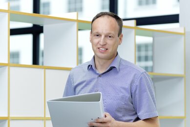 Mann mit dunklen Haaren und blauem Kurzarmhemd steht vor einem Regal mit einem Aktenordner in der Hand. 